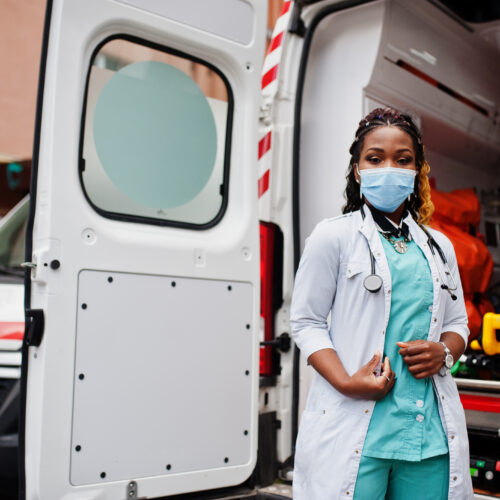 African american female paramedic in face protective medical mask standing in front of ambulance car.